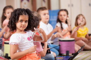 Preschool girl with triangle in focus in front; other children with drum in blurred background