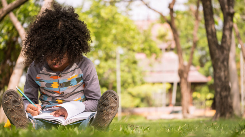 A little boy writing on notebook while sitting on green grass in a park